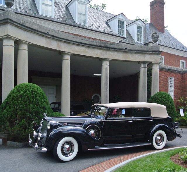Black vintage car parked in front of a brick mansion with white columns and dormers.