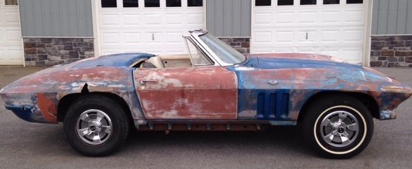 A weathered, blue and red convertible car sits in front of a white garage door.