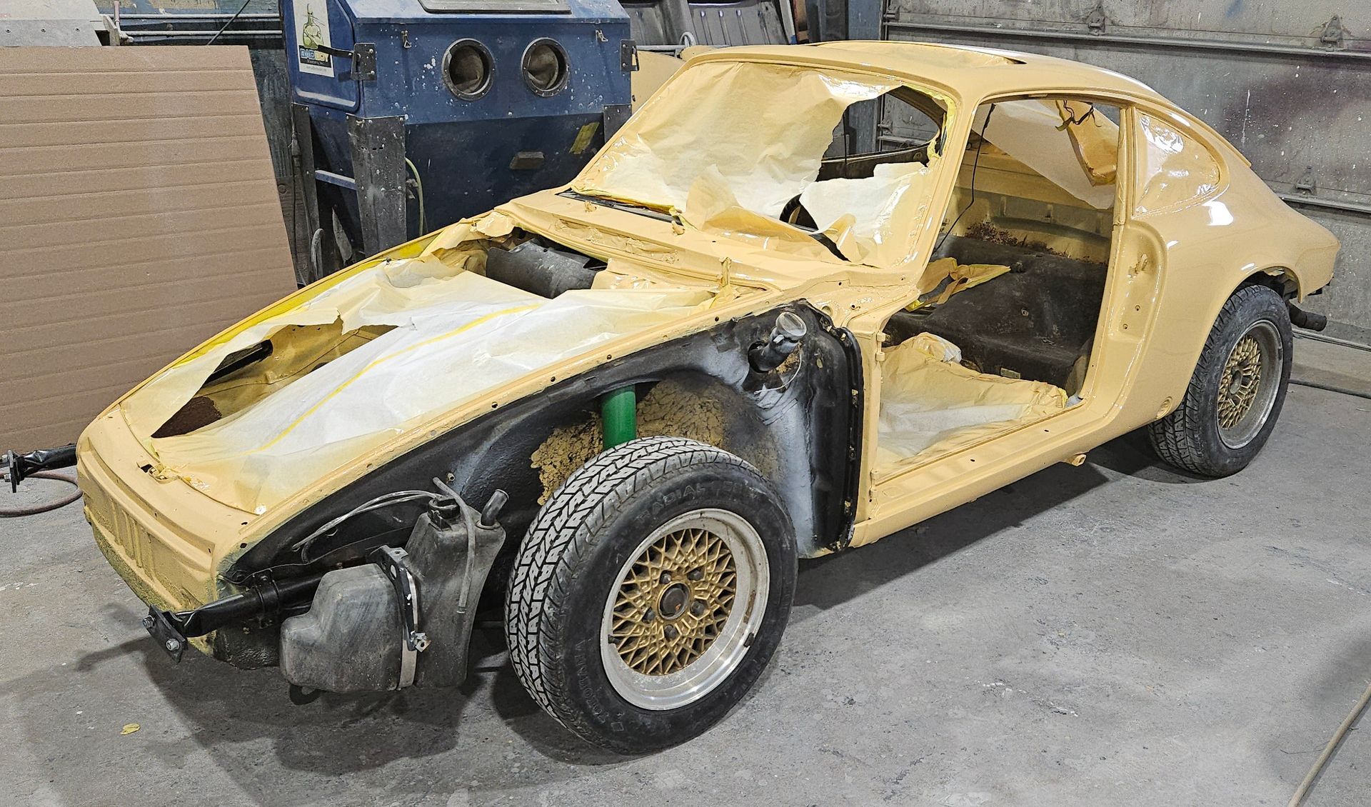 Yellow car body in a workshop, undergoing restoration. Exterior partially stripped, showing suspension and wheels.
