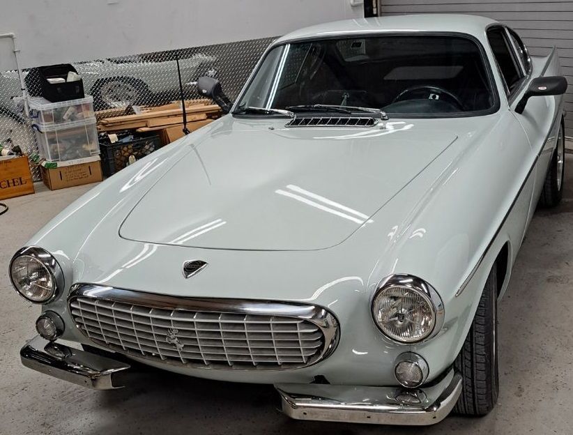 White Volvo P1800 sports car in a garage. Chrome grille and bumpers, round headlights, dark tires.