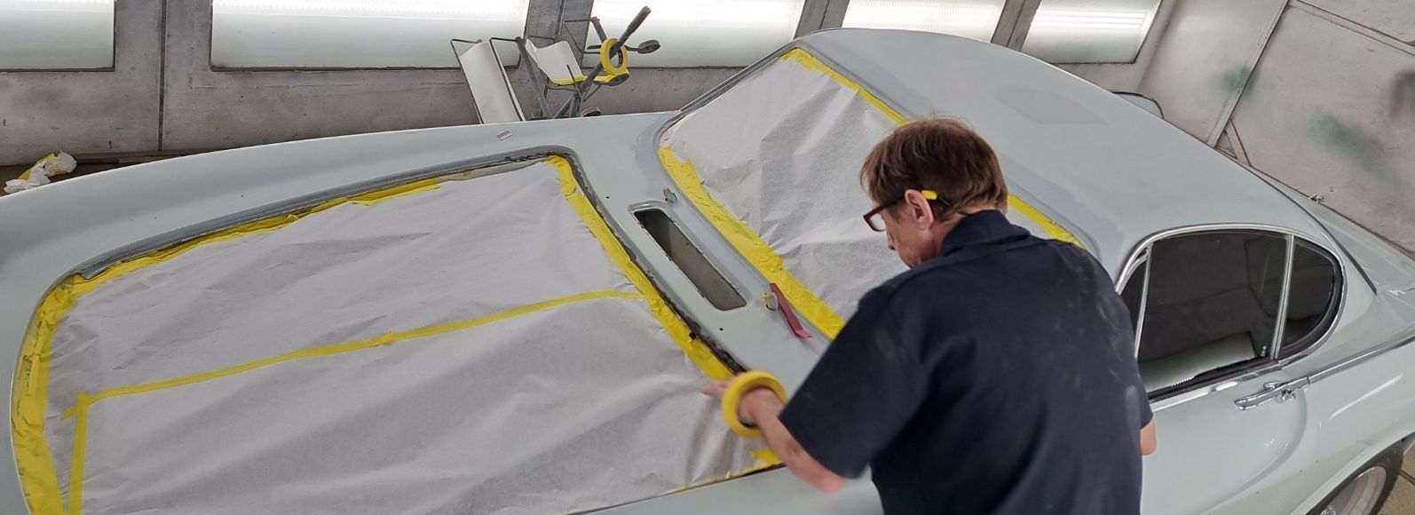 Person masking a light-colored car for painting in a workshop; yellow tape visible.