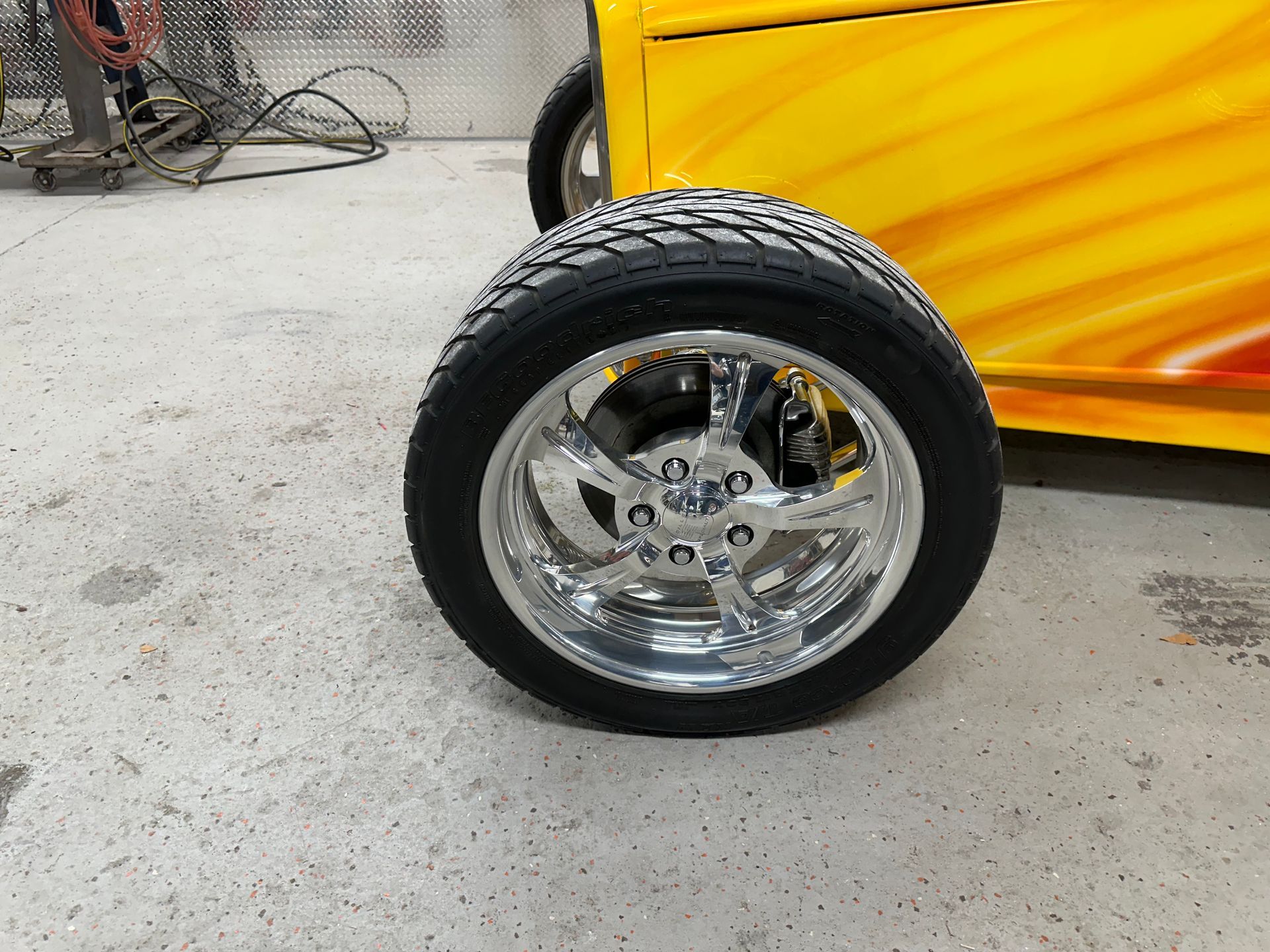 Chrome car wheel and tire against a yellow vehicle with flames; concrete floor.