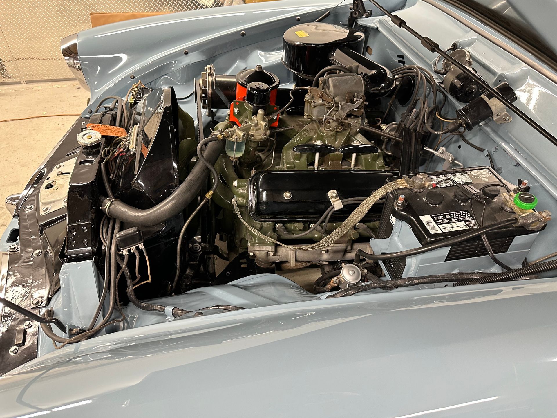 Engine compartment of a classic light blue car, showing the engine, battery, and radiator.