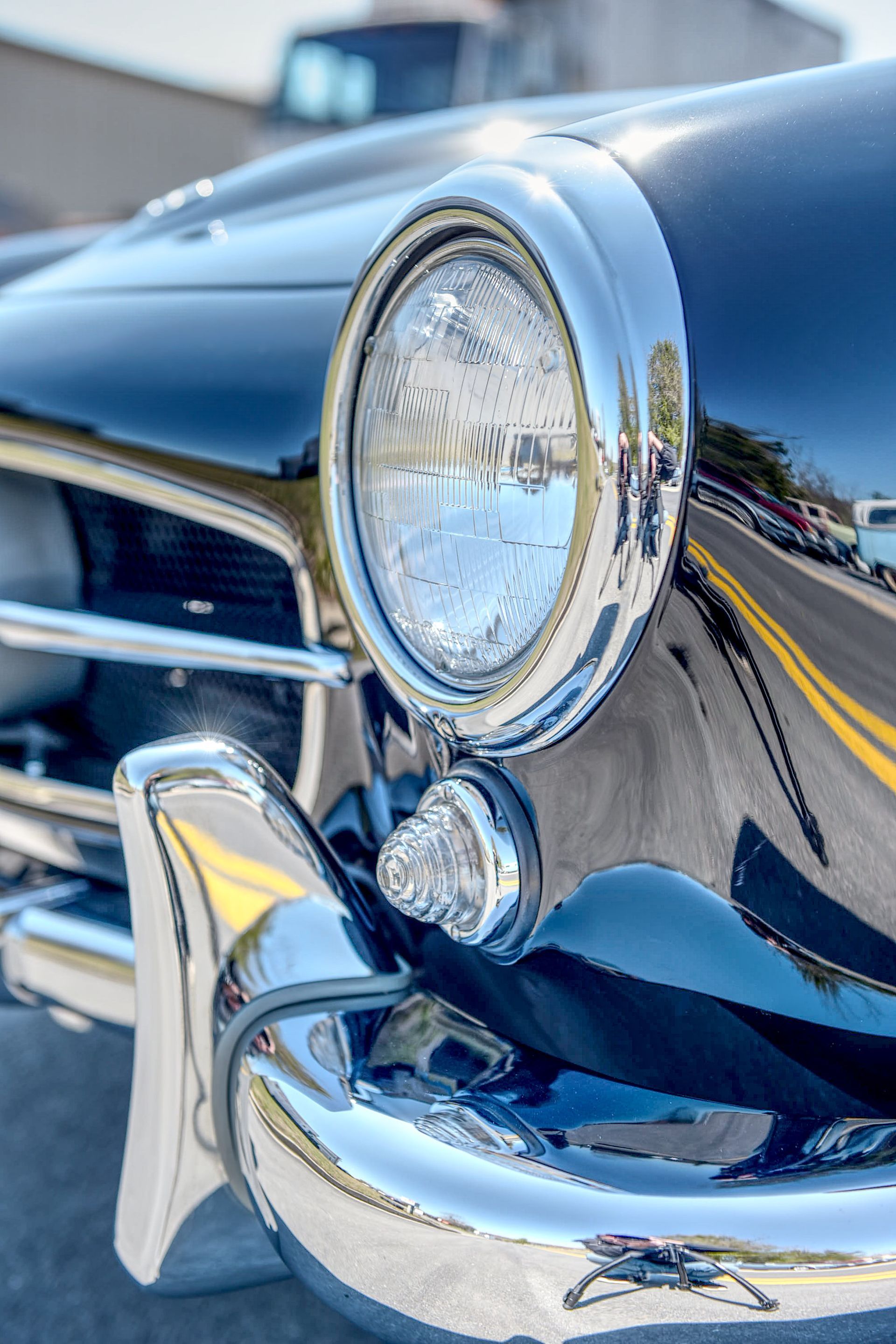 Black classic car headlight with chrome trim, reflecting road and sky.