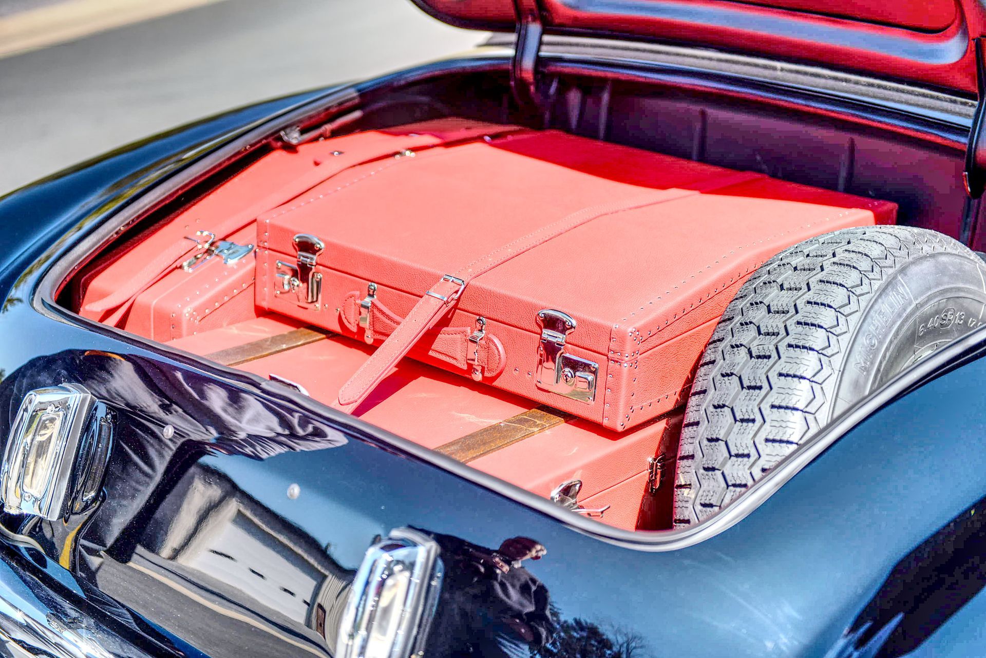 Red luggage in the open trunk of a classic black car with a spare tire.