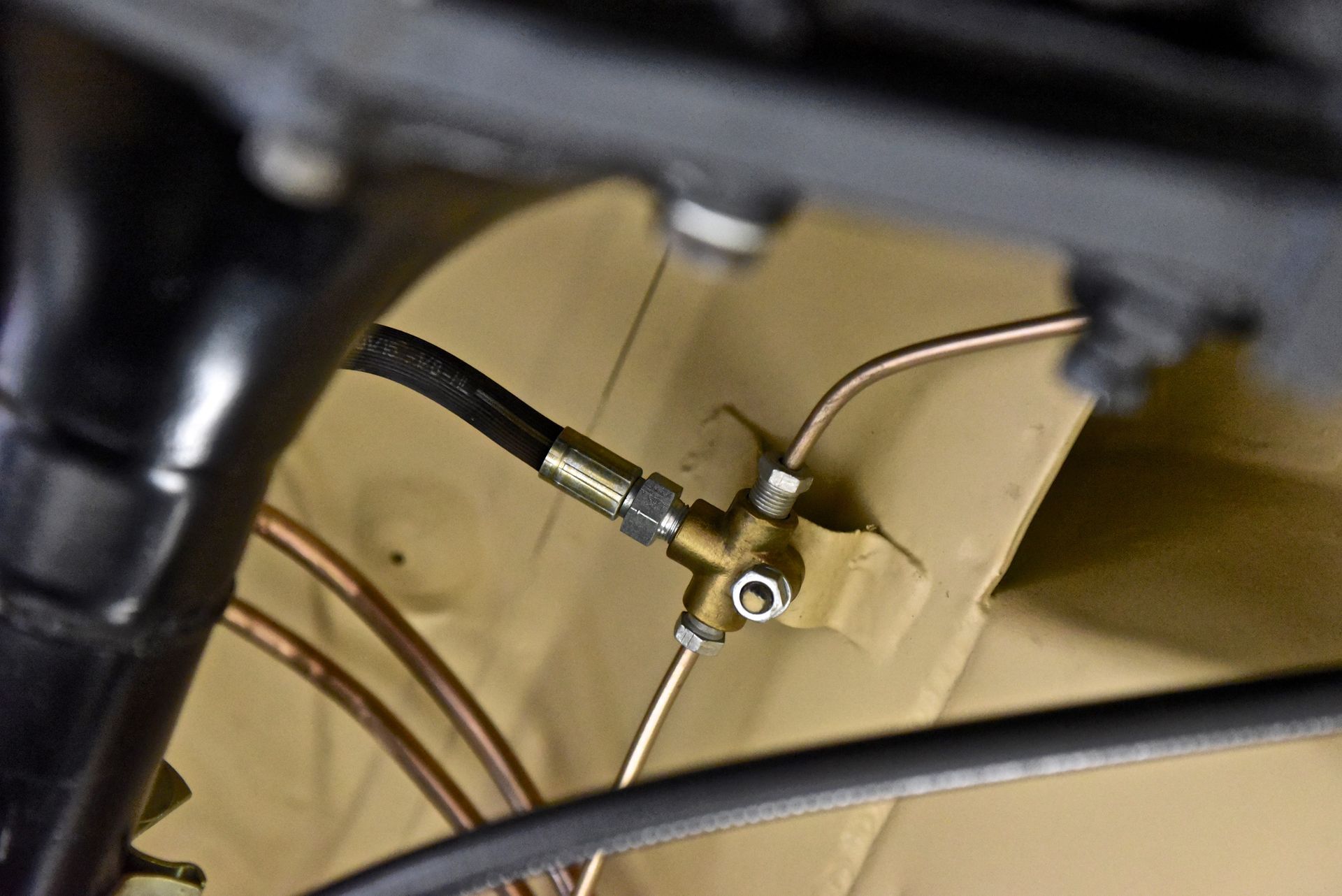 Close-up of car's brake lines. Metal tubing and a black hose connect to a brass fitting on a tan surface.