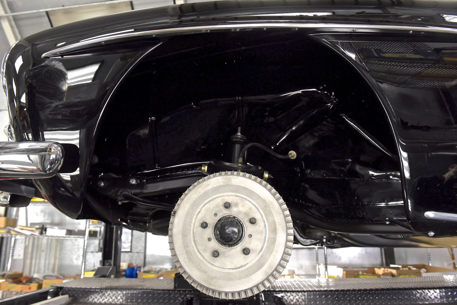 Black car's rear wheel and suspension components viewed from below, undergoing restoration in a shop.