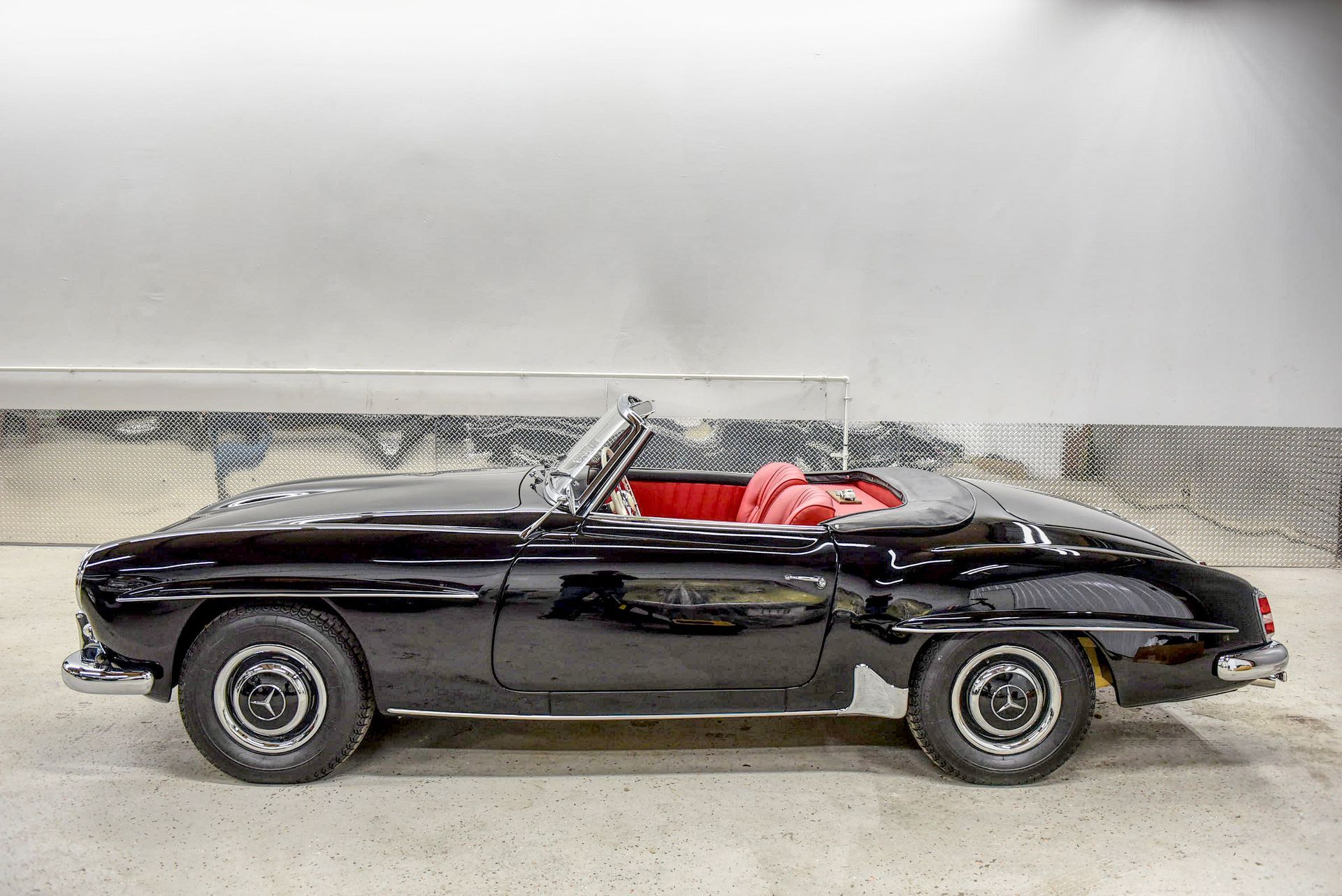 Black classic Mercedes-Benz 190 SL convertible with red interior, parked indoors.