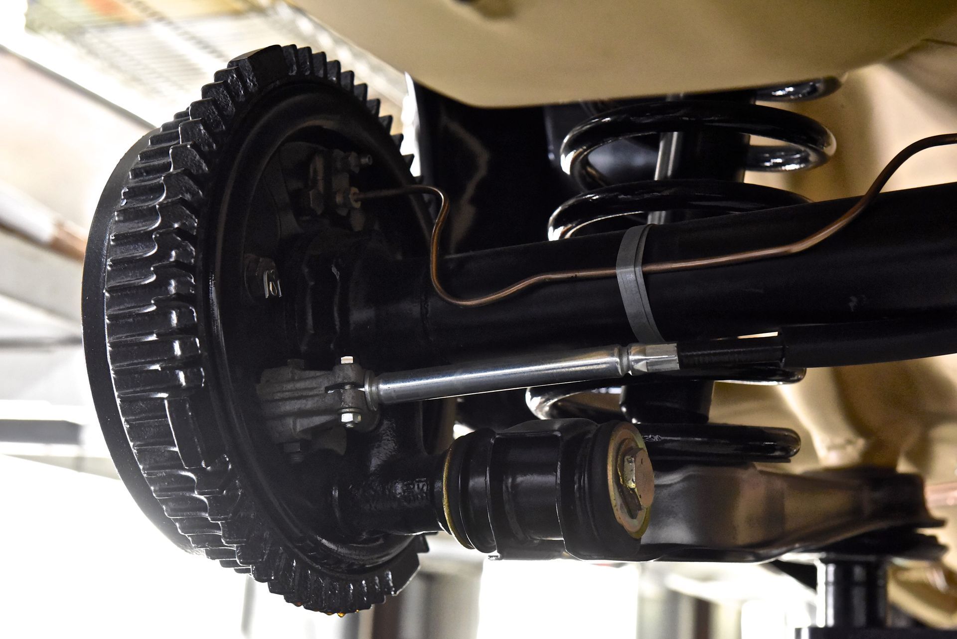 Black car rear axle with drum brake, coil spring, and suspension components, viewed from below.