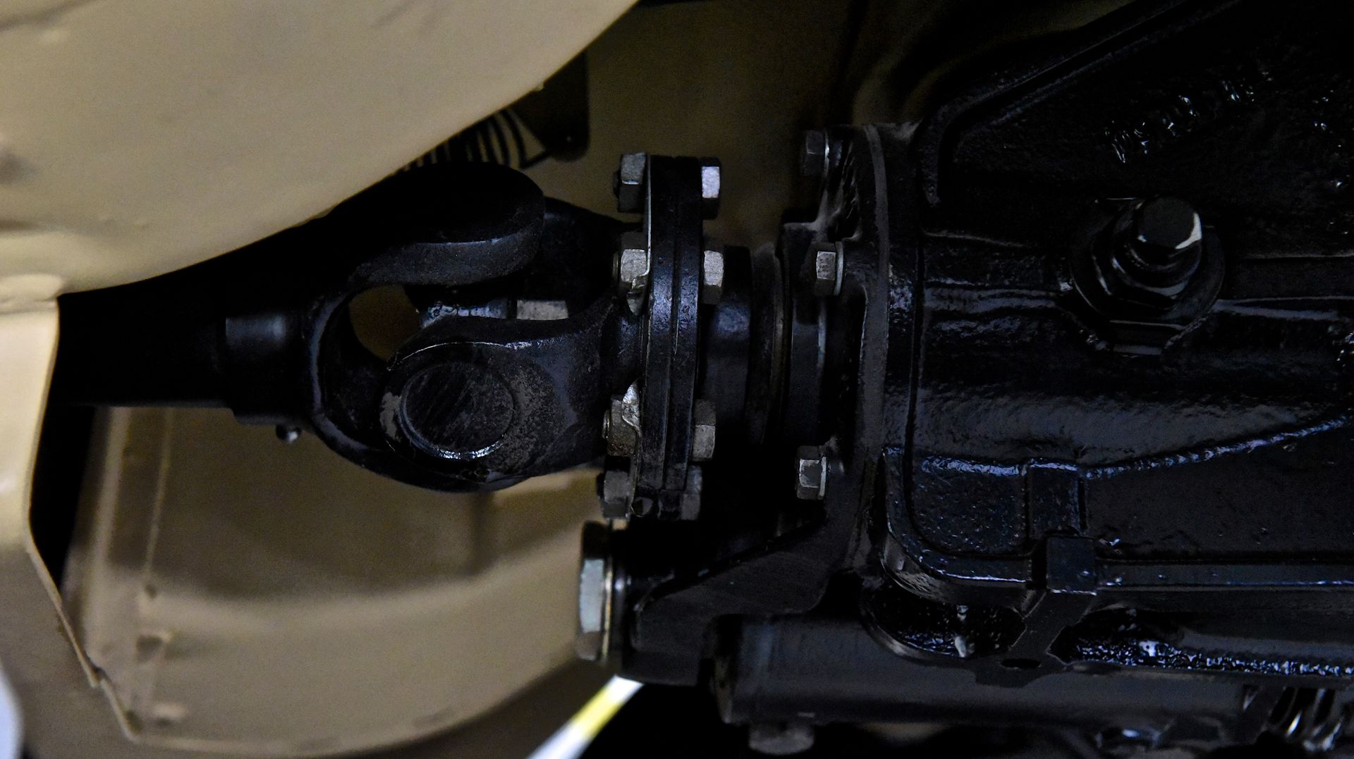Black driveshaft connecting to a black transmission with bolts, seen from underneath the beige car frame.
