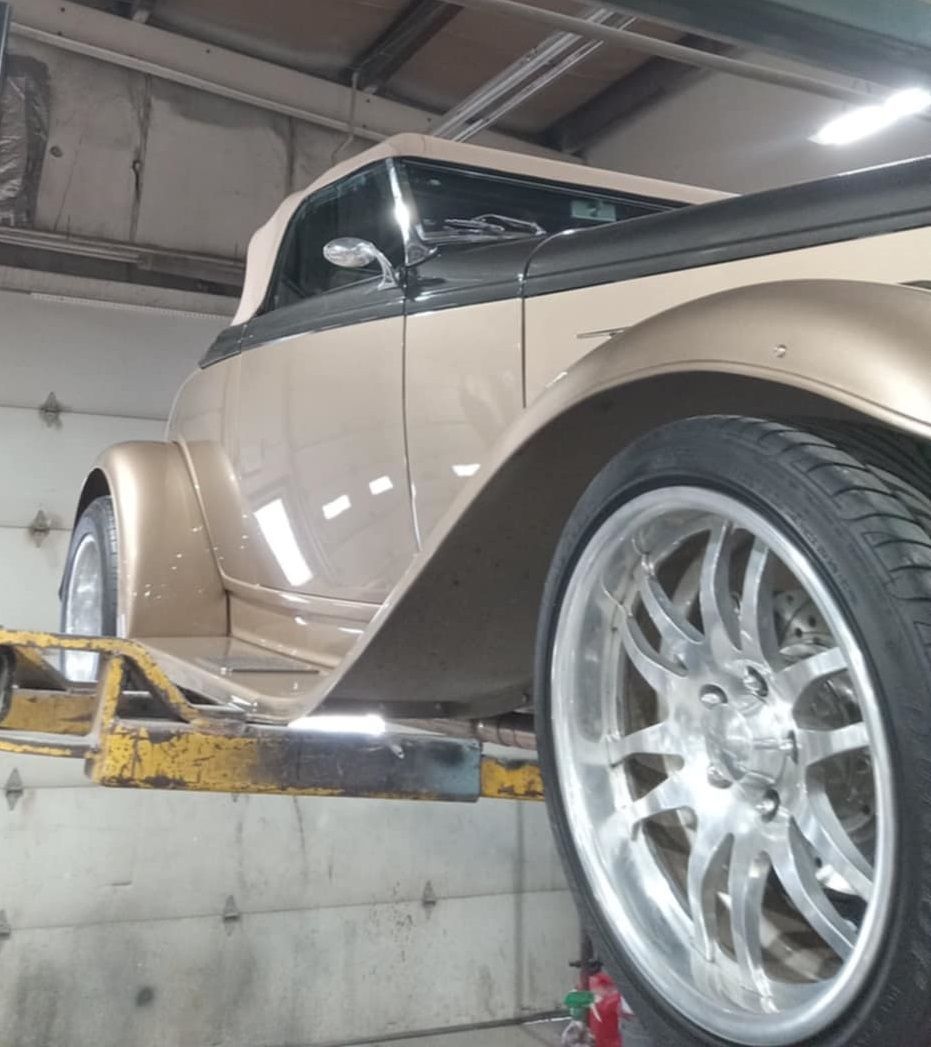 Gold classic car raised on a lift; chrome wheels, beige body, black top, in a garage.