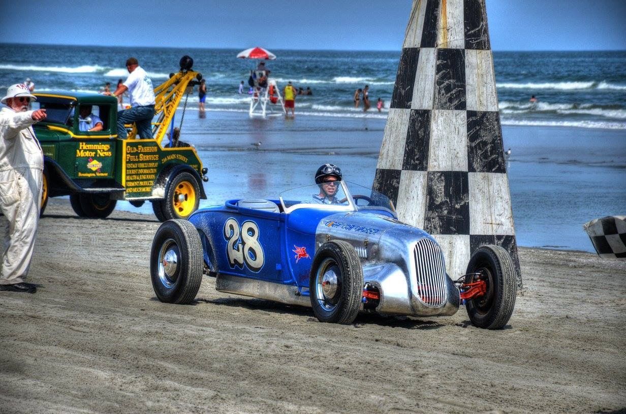 Blue race car #28 on a beach, near a checkered flag, people in the background.