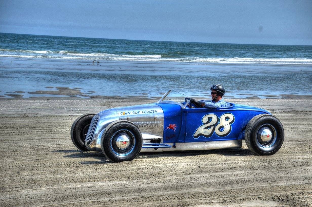 Hot rod on beach, blue and silver, #28 on side. Driver wearing helmet. Ocean in background.