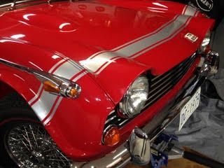 Red vintage car with silver racing stripes on the hood and fender, parked indoors.