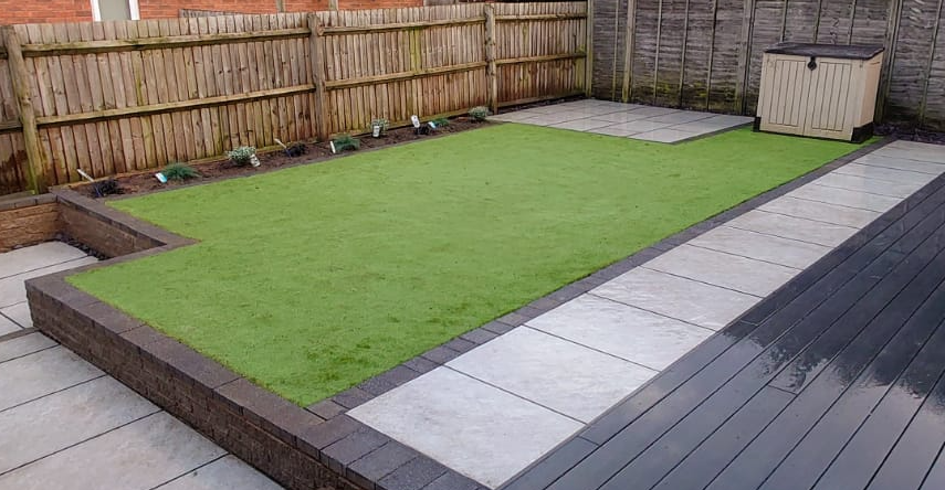 Raised artificial lawn edged in dark brick beside porcelain paving and dark composite decking