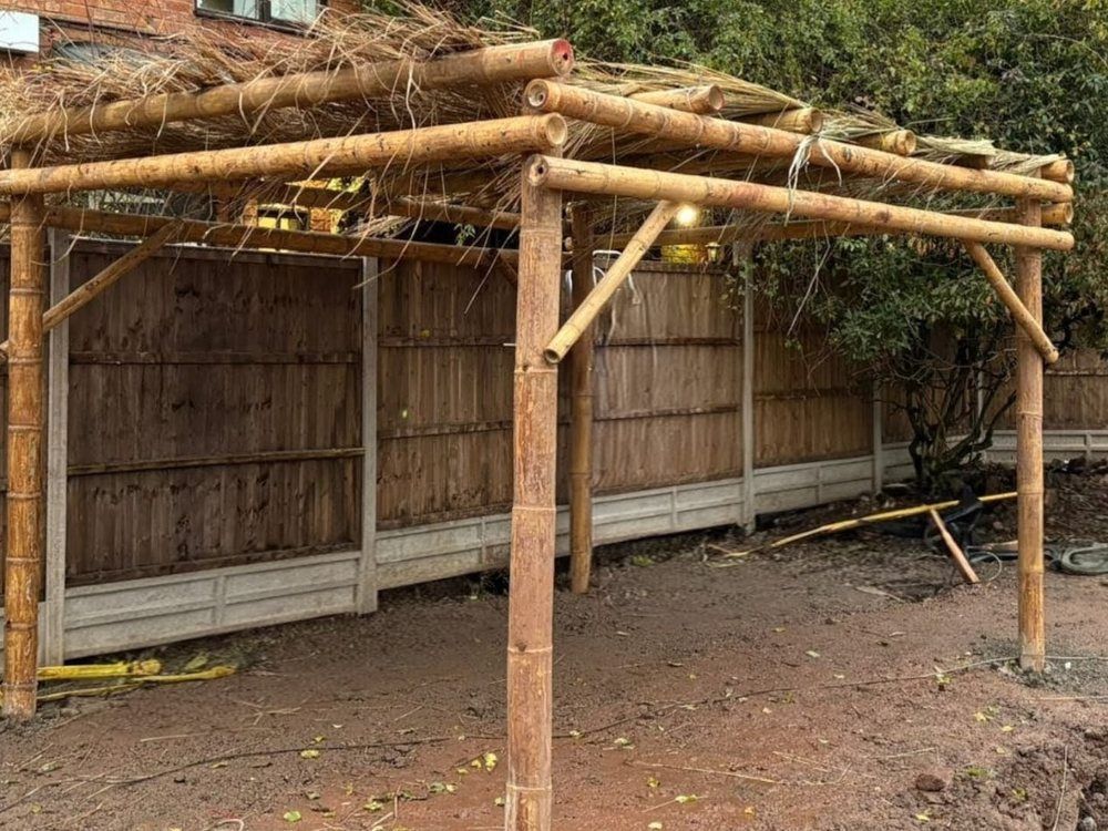 Bamboo-framed pergola with a thatch-style roof being built on bare ground beside a close-boarded fence