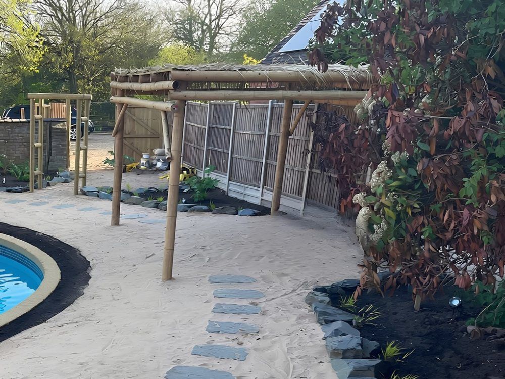 Bamboo-framed pergola with a thatch-style roof over beach sand, with slate stepping stones, slate-edged planting and a timber ladder frame