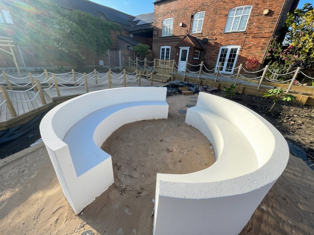 Two curved white rendered bench seats forming a circle on beach sand, with the rope-edged boardwalk and brick farmhouse behind