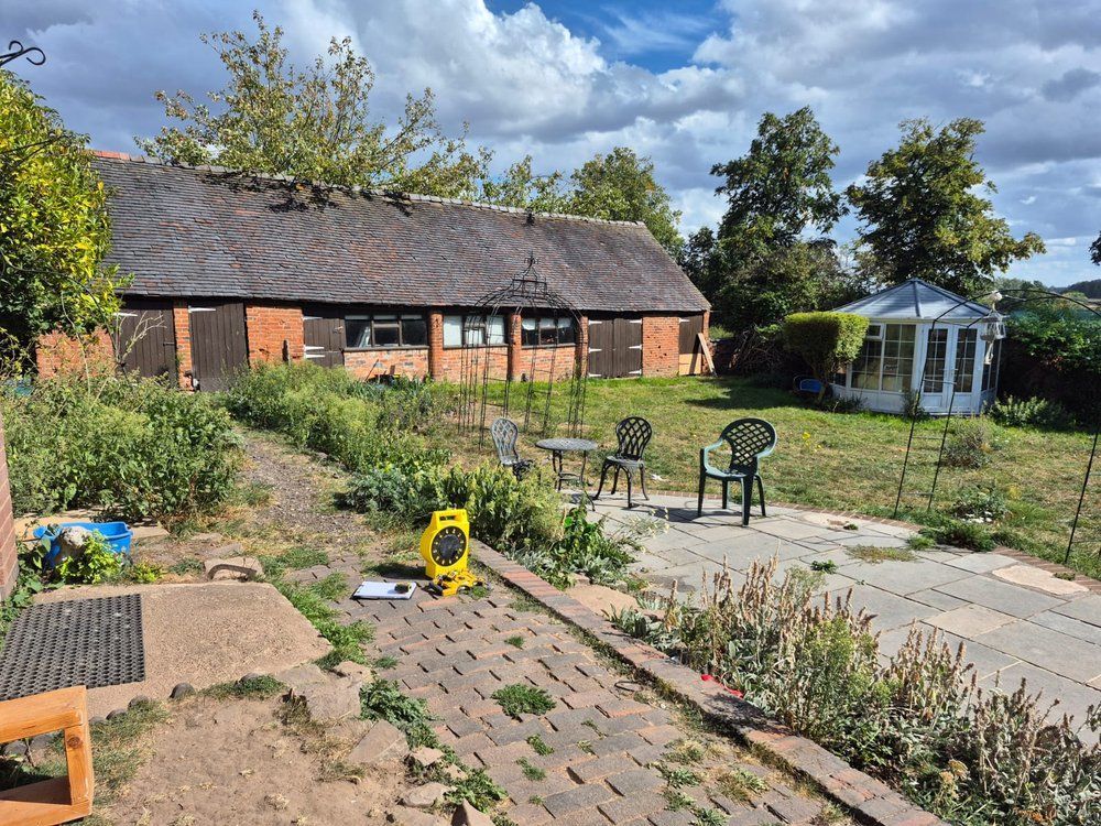 Overgrown borders, a weathered brick path and a paved patio in front of a brick barn at Top Leather Mill Farm, Atherstone, before landscaping