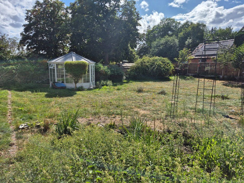 Large open lawn with patchy grass, a white summerhouse and metal garden arches before the tropical garden was built