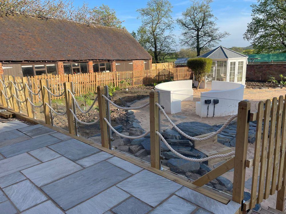Grey stone patio with a glass and rope balustrade and picket gate, overlooking slate rocks, the curved fire pit seating and a white summerhouse