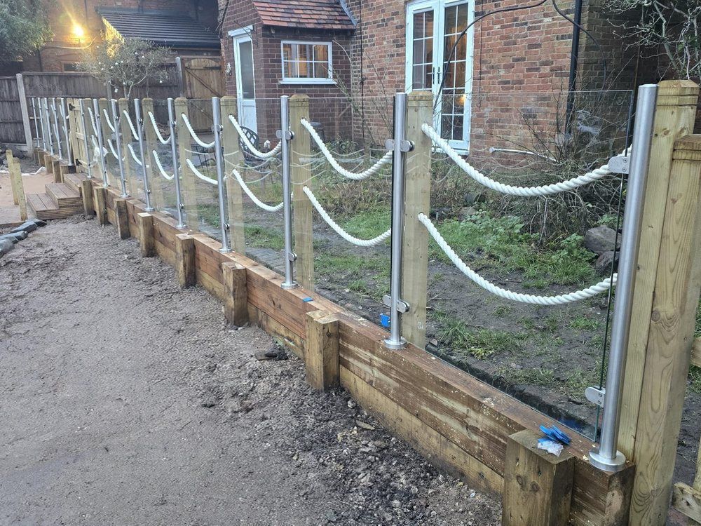 Glass balustrade panels with stainless steel posts and rope detailing on a timber sleeper retaining wall beside a brick farmhouse