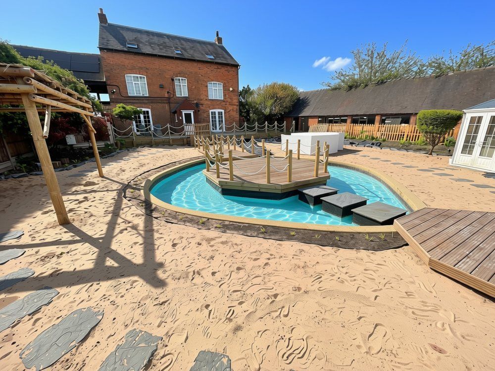 Turquoise lagoon with a rope-edged octagonal island deck and three stepping blocks leading to a small deck, surrounded by beach sand at Top Leather Mill Farm, Atherstone
