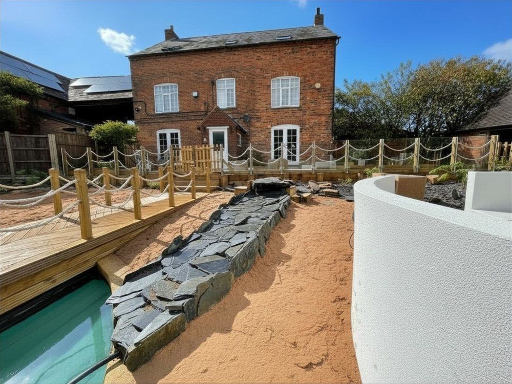 Slate-lined waterfall channel running across sand from the lagoon towards a brick farmhouse with a glass and rope balustrade