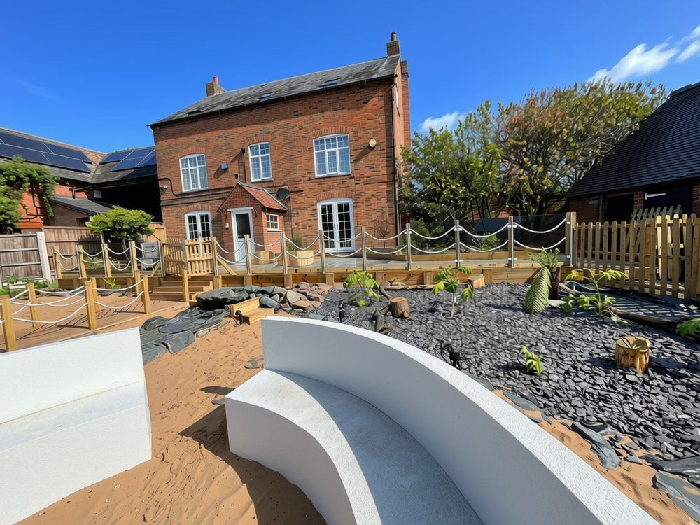 Young tropical-style plants and ferns in a bed mulched with slate chippings beside curved rendered seating, with the farmhouse behind