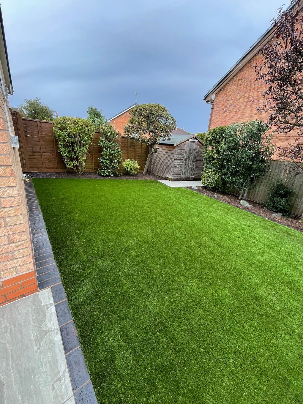 Artificial lawn running from a paved border to shrubs and fencing at the end of a garden