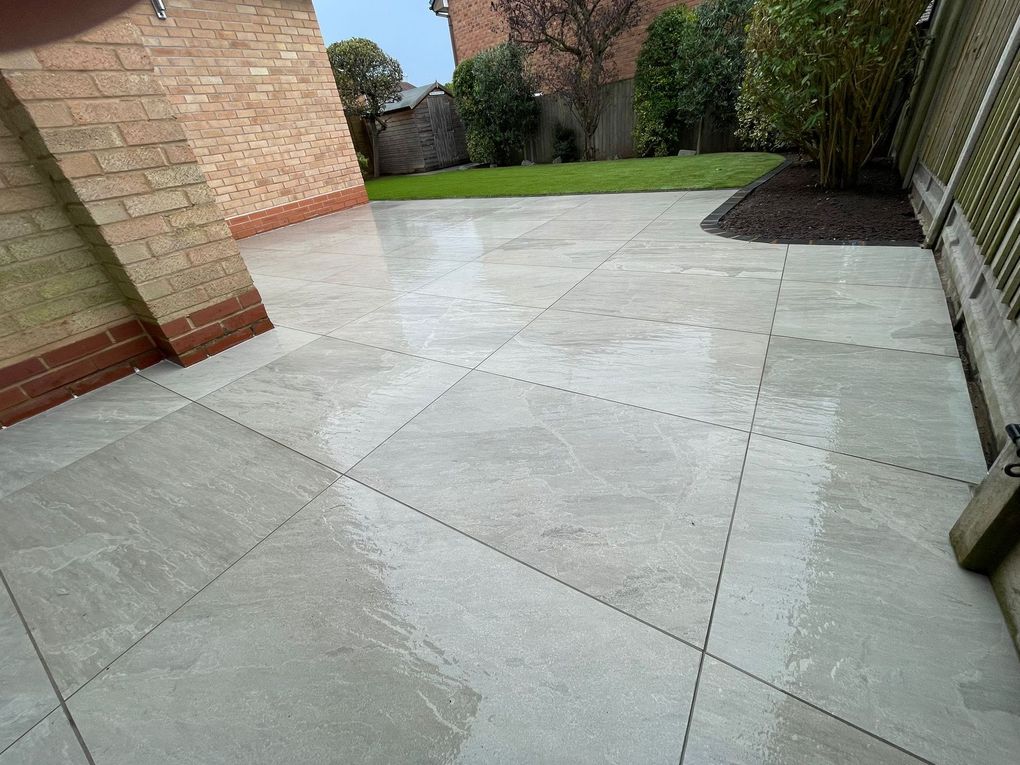 Large-format light grey porcelain patio laid beside a brick house with a lawn beyond