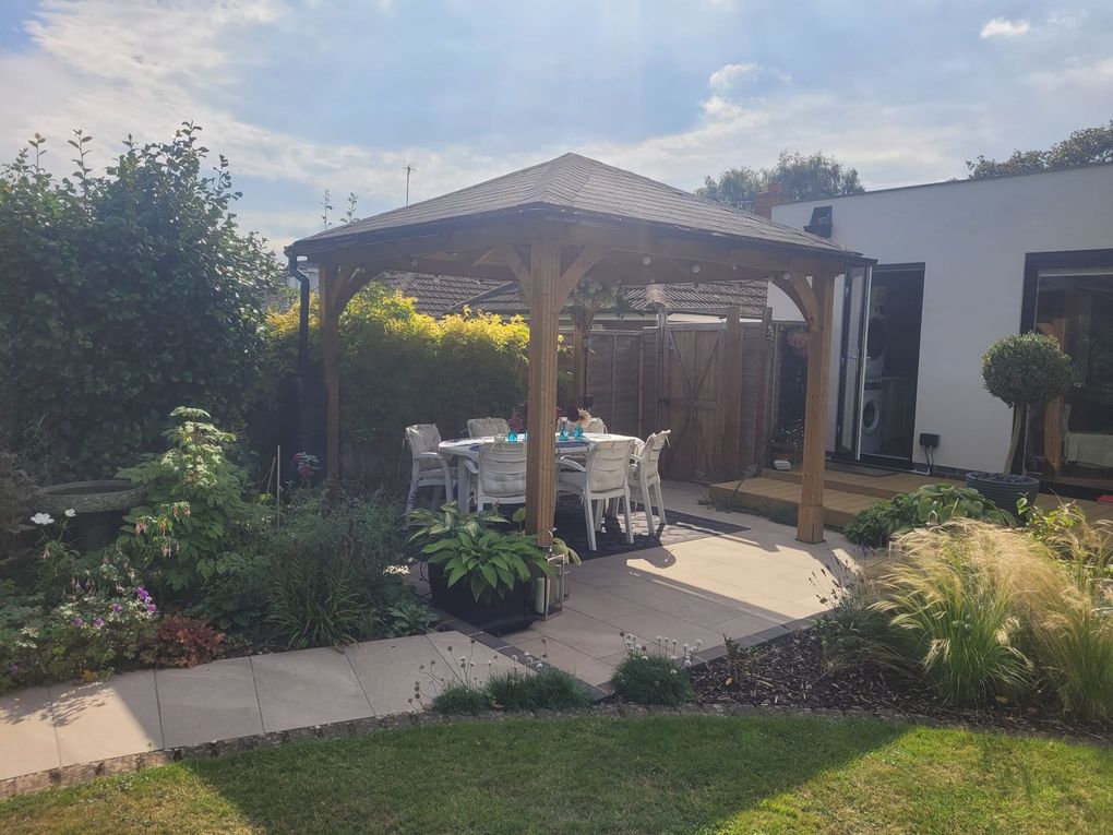 Timber gazebo with a tiled roof over a dining table on a paved terrace, surrounded by planting