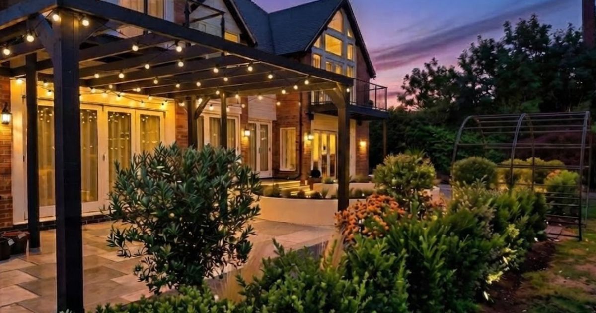 Warm-lit patio garden beside a glass house at dusk, with lush plants and a purple sunset sky.