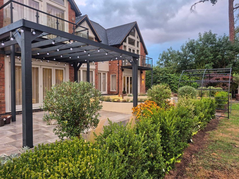 Dark open-top timber gazebo with festoon lighting beside established planting and clipped evergreens at Lapworth