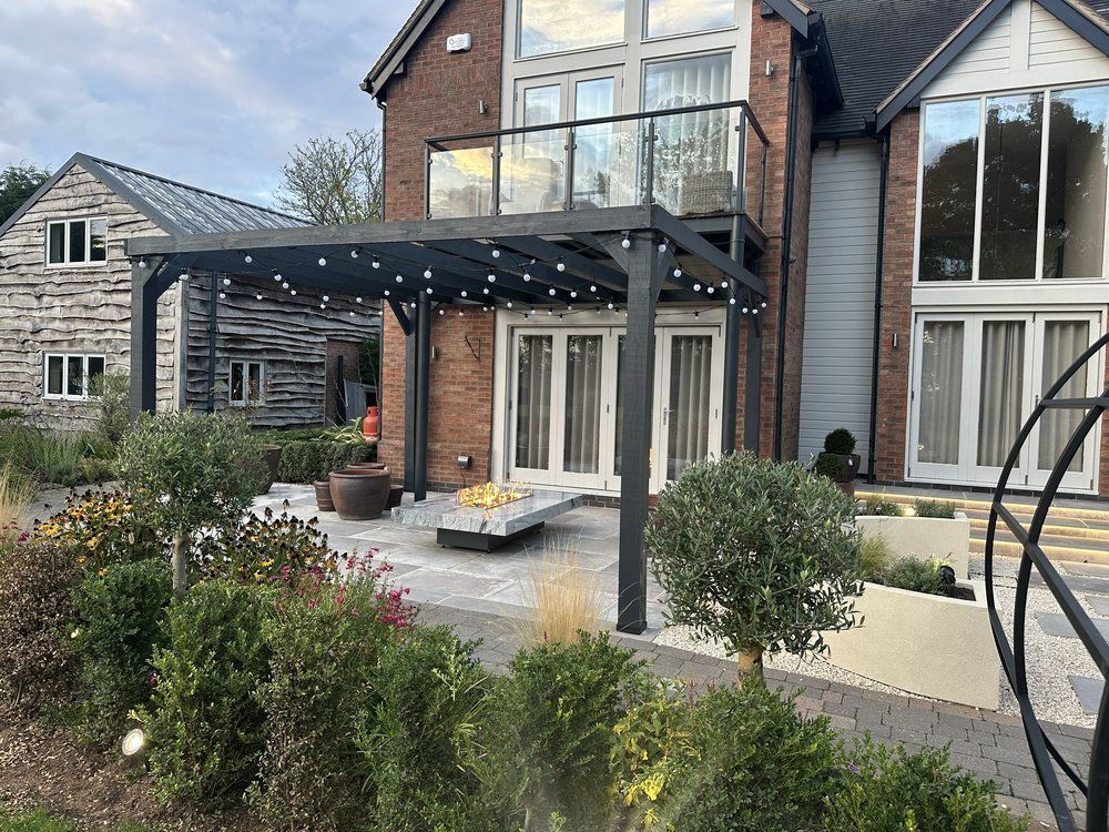 Timber gazebo with festoon lighting over a seating area with a fire table, framed by planting and a Moon Gate at Lapworth