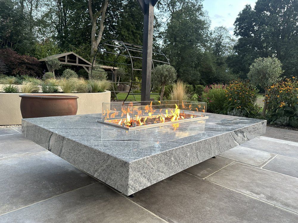Lit granite fire table on porcelain paving with ornamental grasses and trees behind at Chestnut House, Lapworth