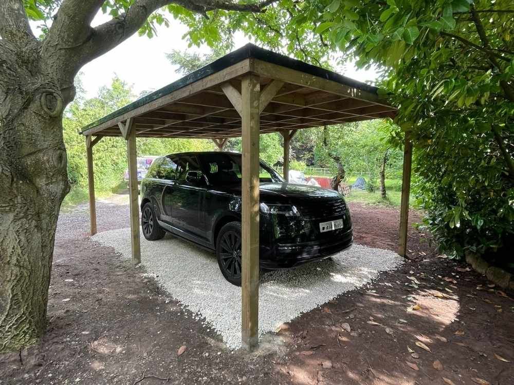 Open-sided timber-framed carport on a gravel base beneath mature trees at Quarry House near Coleshill