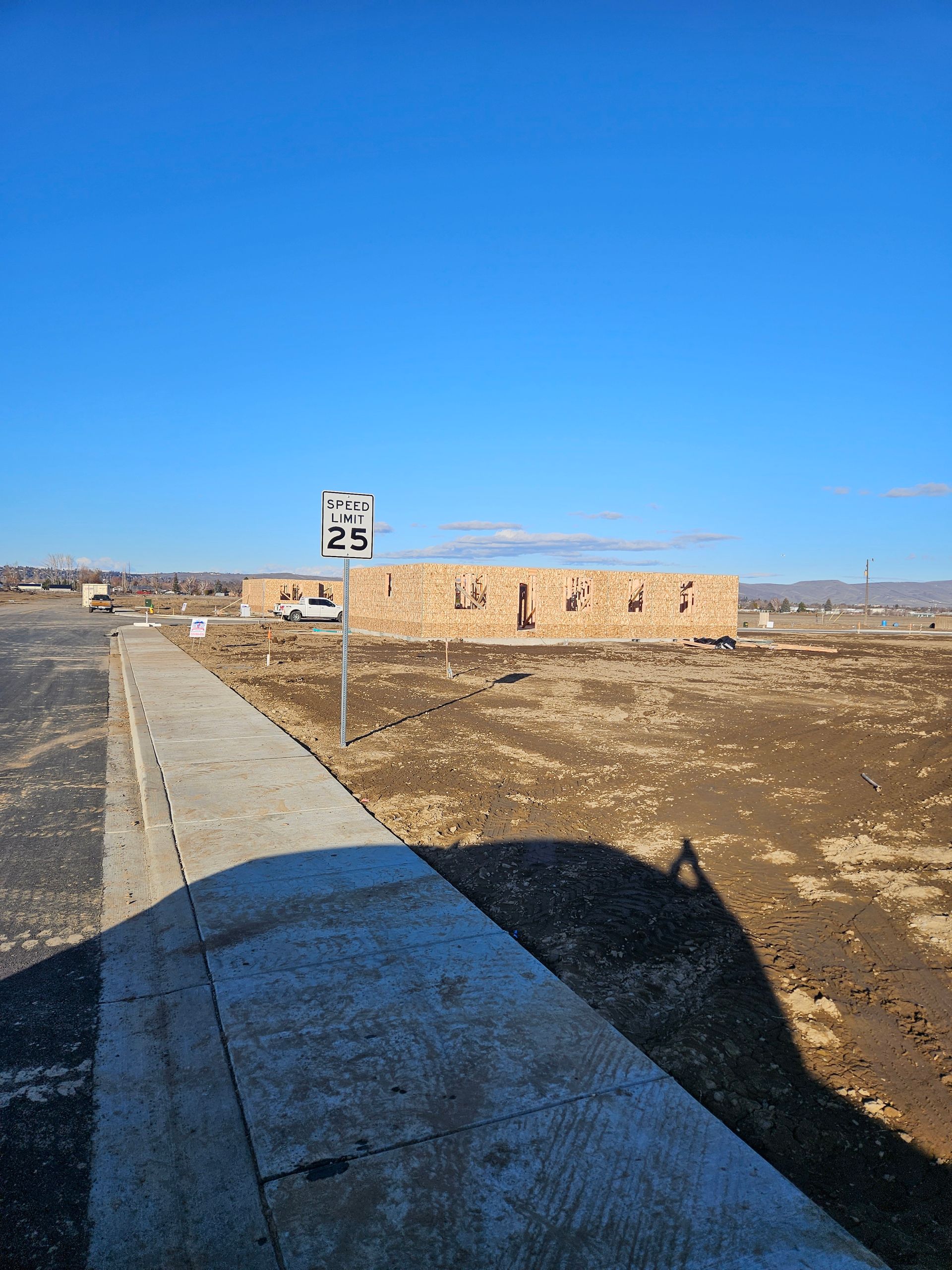Empty dirt lot with a low brick building under a clear blue sky, viewed from a sidewalk.