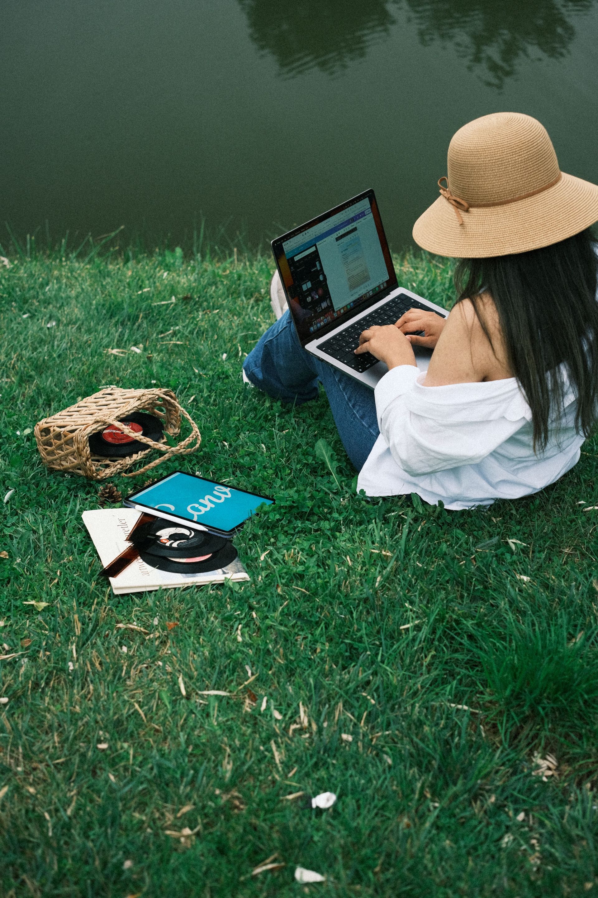 Person in a straw hat sits on grass by water, using a laptop beside a bag and books.