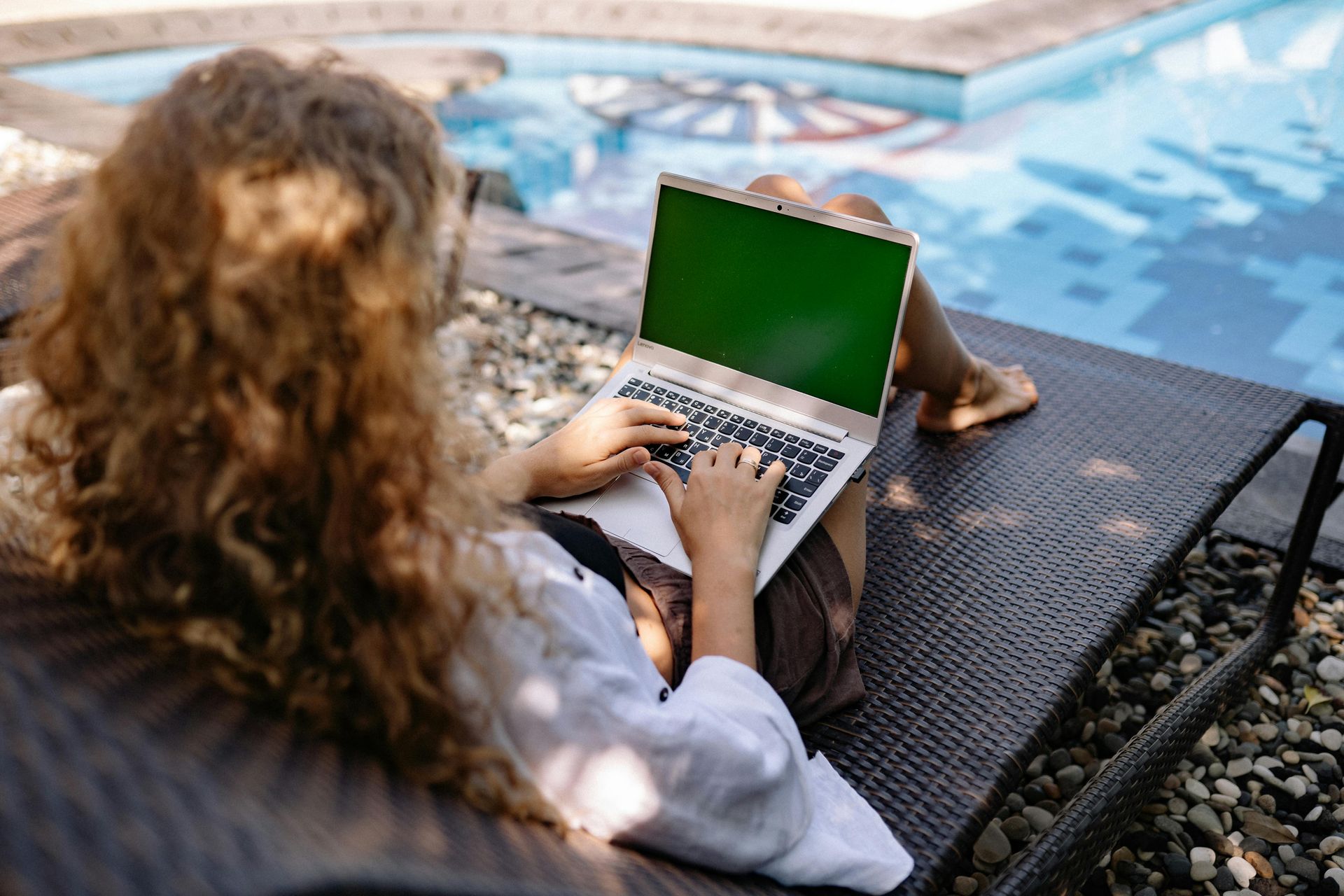 Person typing on a laptop beside a pool, with a green screen and a bag on the chair nearby.