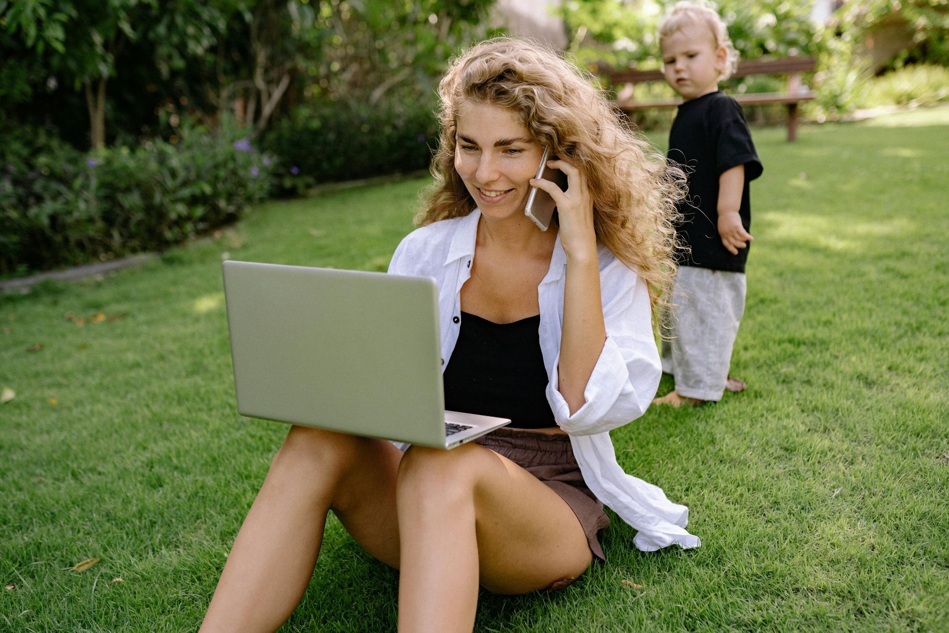Woman using a laptop on grass while a child stands nearby in a sunny park