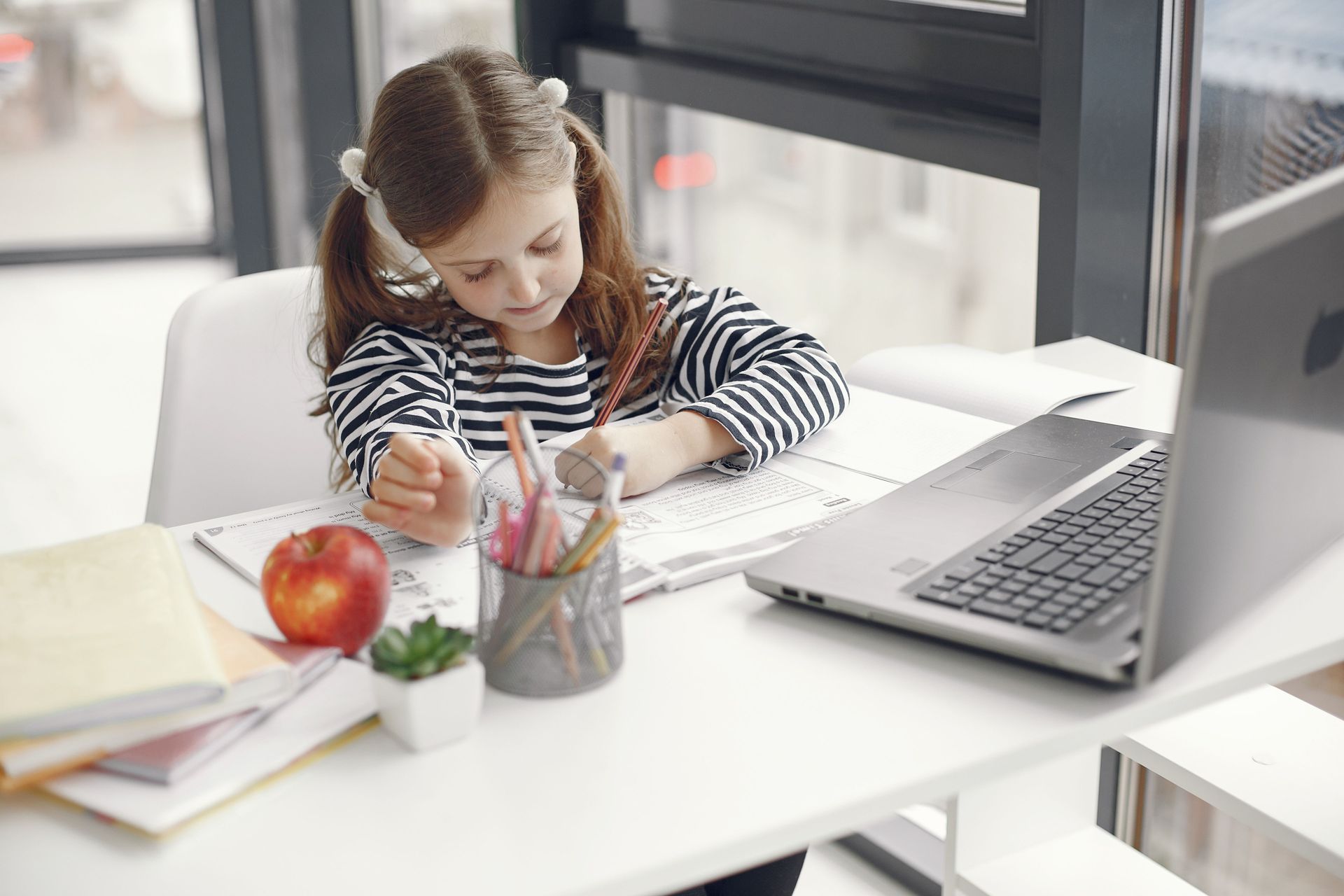 Child writing at a desk beside a laptop, with an apple and cup of pencils nearby