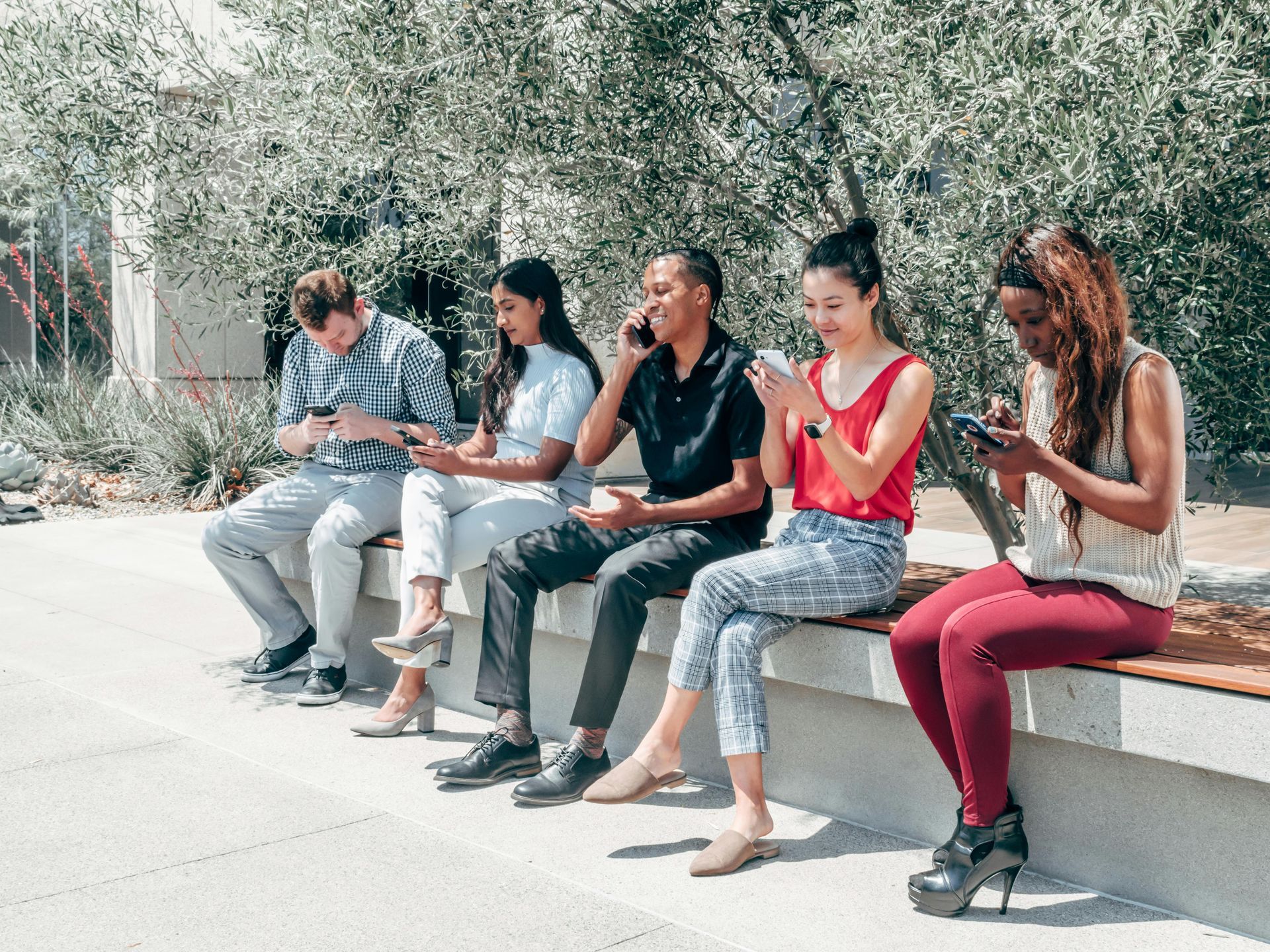 Five people sit on a low wall outdoors, chatting and using their phones.