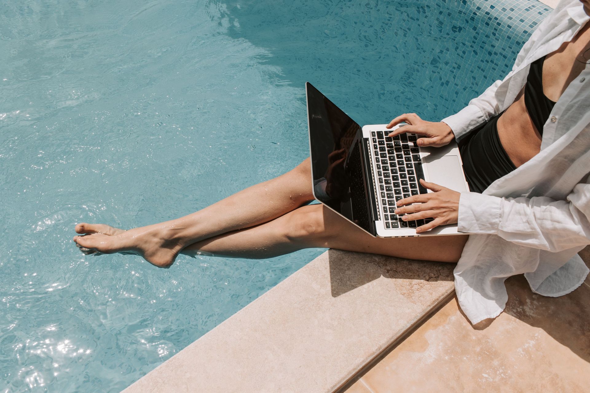Person using a laptop while sitting by a turquoise pool, legs extended into the water