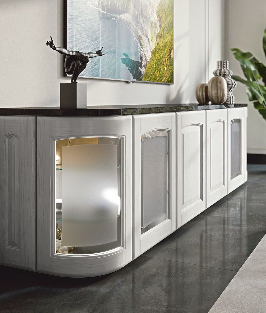 Mobile bianco con ante in vetro, decorato con opere d'arte e decorazioni, contro una parete con un dipinto paesaggistico.