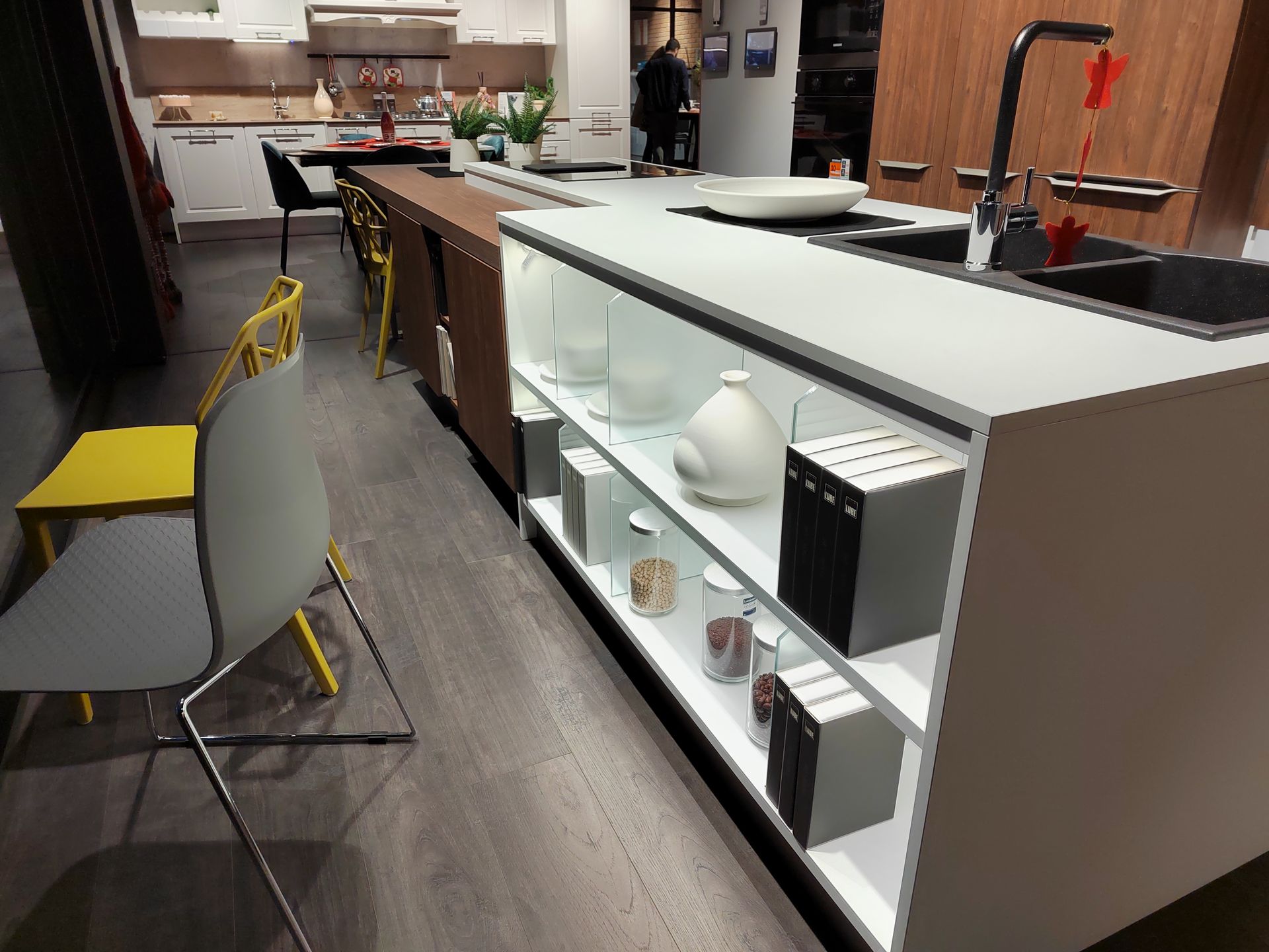 Showroom di cucine con una lunga isola. Mobili moderni, piano di lavoro bianco e vari articoli da cucina in esposizione.