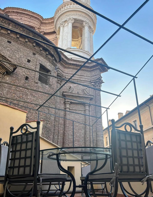 Tavolini da caffè all'aperto con sedie e tavolo in metallo nero, protetti da una rete, accanto a un vecchio campanile in mattoni, sotto un cielo azzurro.