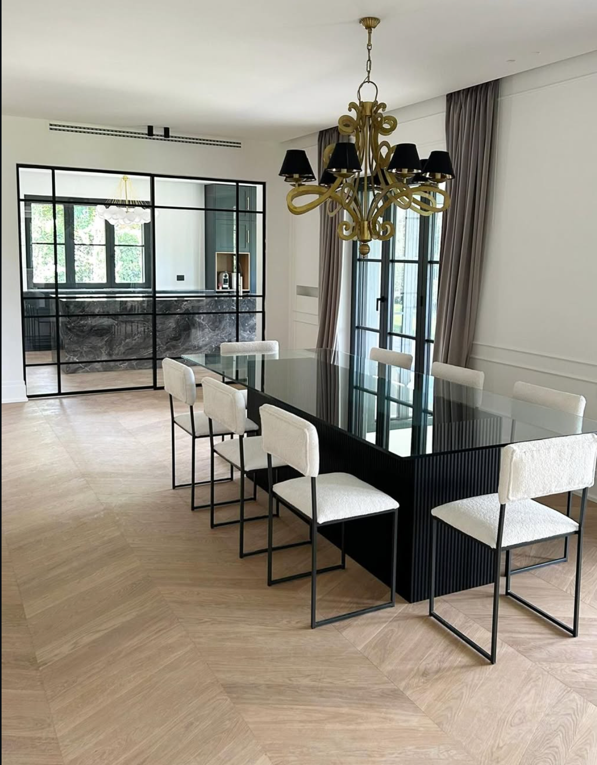 Sala da pranzo moderna con un lungo tavolo di vetro, sedie bianche, lampadario dorato e finestre con cornici nere.