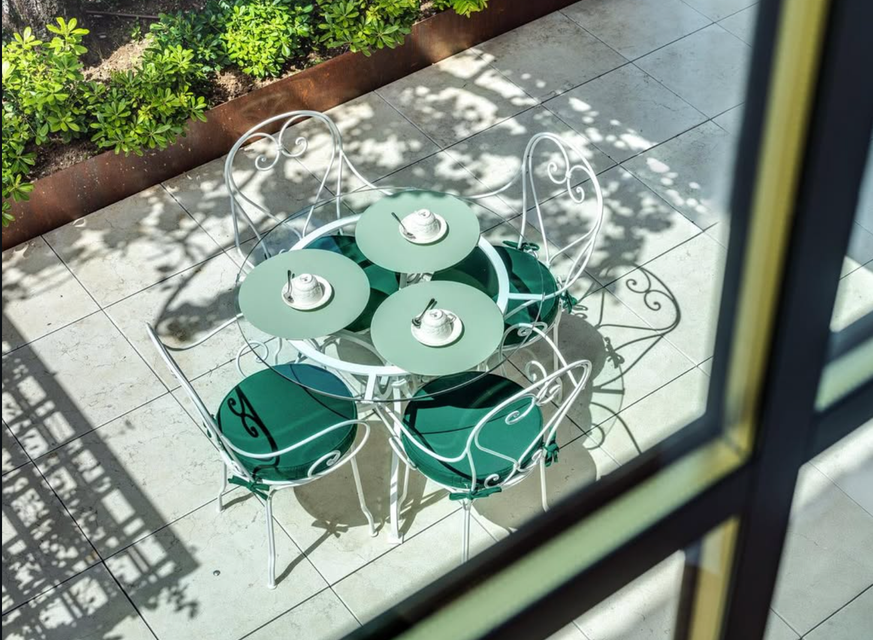 Tavolino da caffè all'aperto con quattro sedie verdi, visto attraverso una finestra, con due tazze sul tavolo e la luce del sole che filtra tra gli alberi.