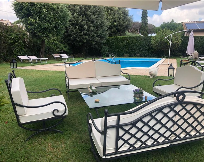 Salotto da patio allestito sul prato a bordo piscina, con sedie imbottite bianche e tavolo in vetro.